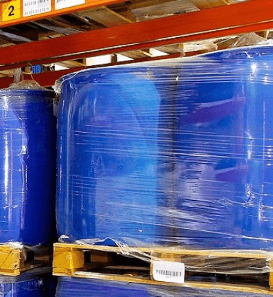 Stacks of large blue barrels wrapped in plastic sit on wooden pallets in a warehouse.