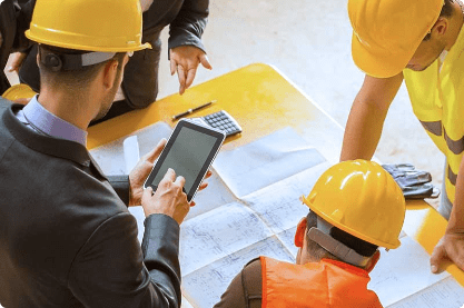 Group of construction workers in hard hats and a suited man with a tablet, examining blueprints on a table, conveying teamwork and planning.