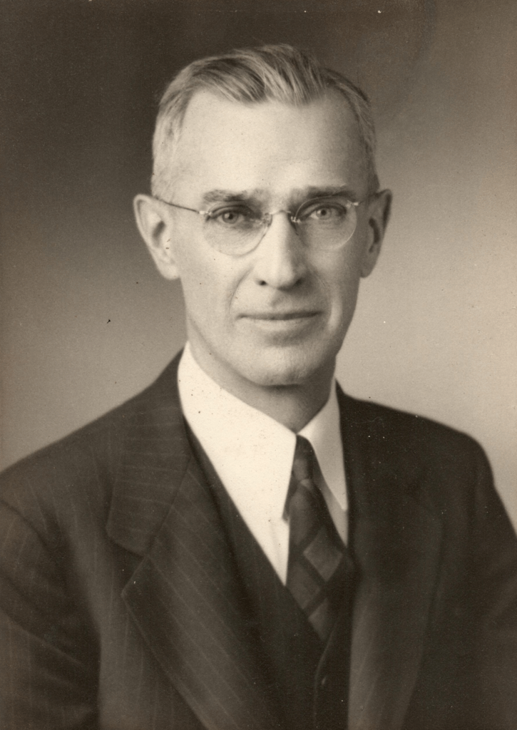 Black and white portrait of an older man with glasses and a serious expression, wearing a suit and tie.