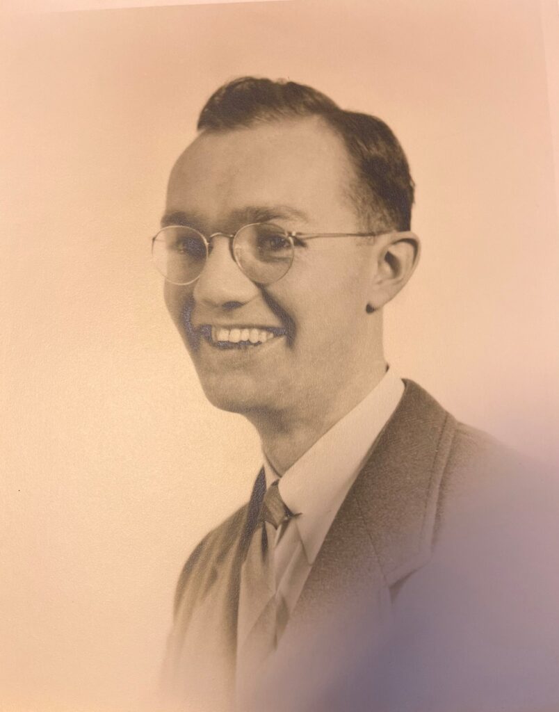 Black and white portrait of a smiling man with glasses, wearing a suit and tie.